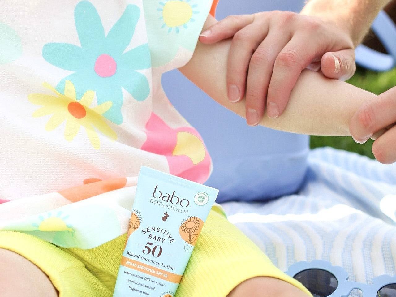 A child holds Sensitive Baby sunscreen while lotion is applied outside.