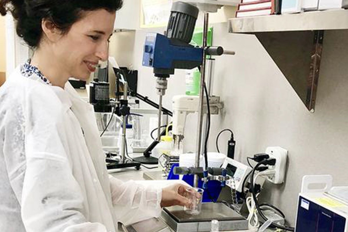A woman in a lab coat handles a beaker beside mixing equipment.