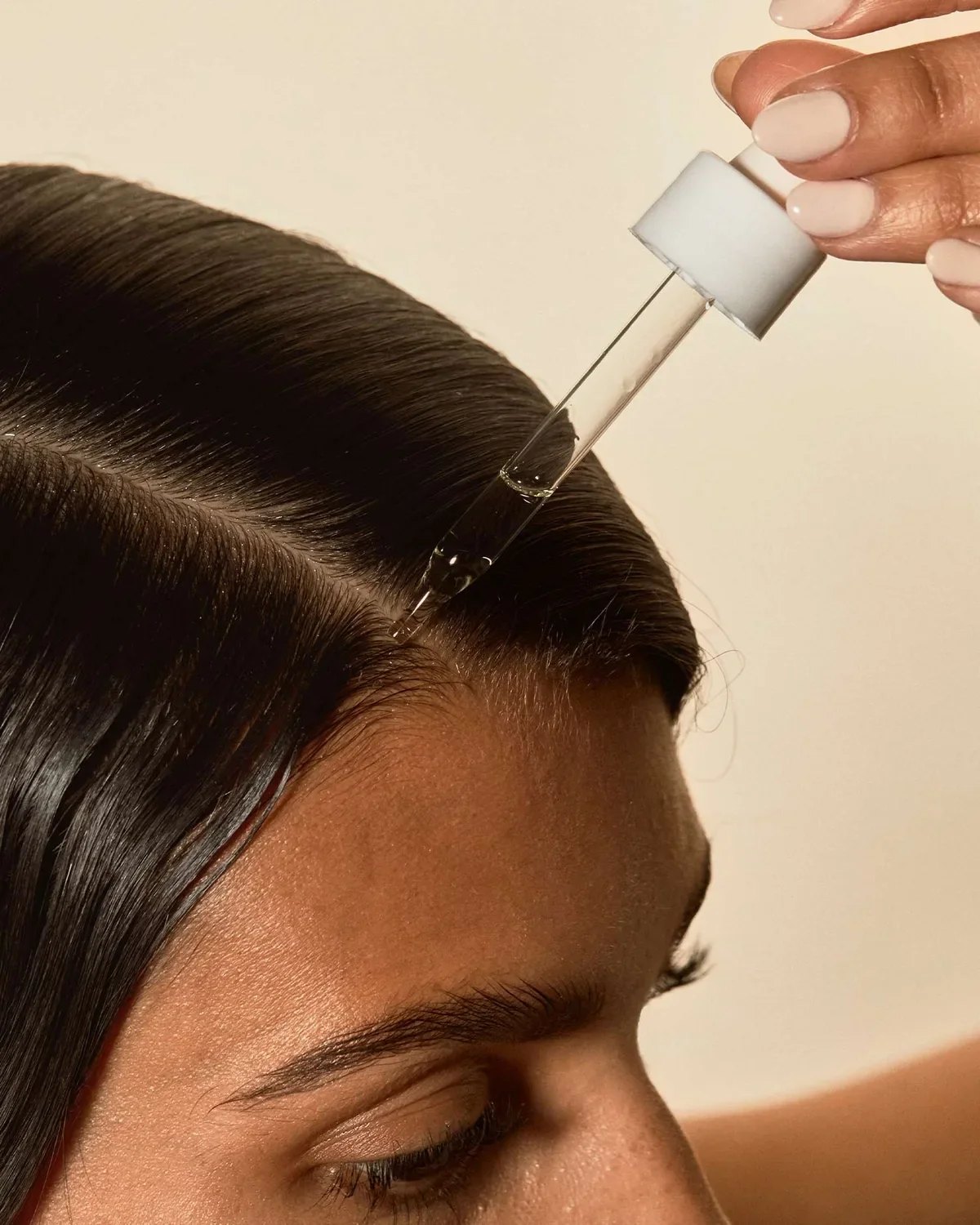 Dropper applies scalp oil to parted dark hair closeup.